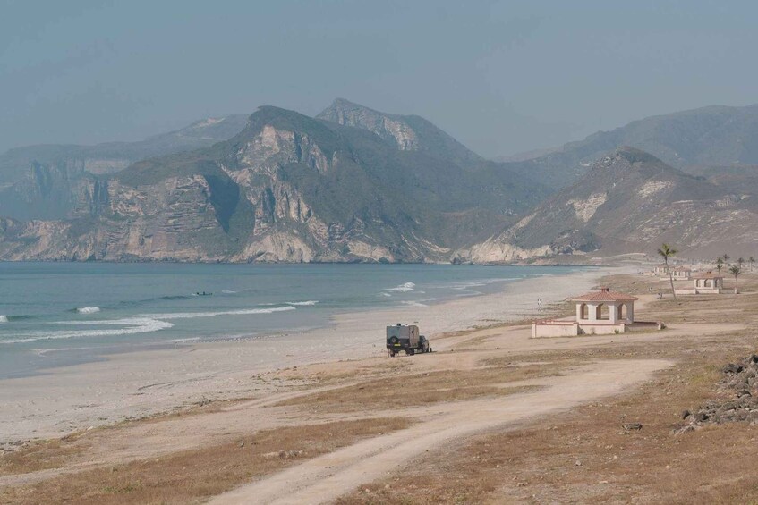 A coastal landscape with a dirt road, a small vehicle, and a few buildings near the beach. 2