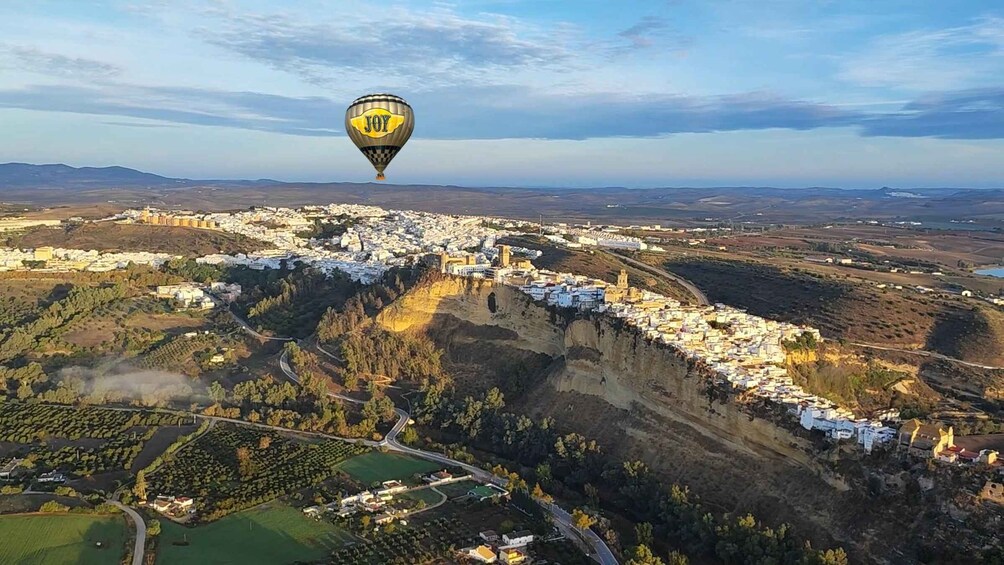 A hot air balloon over a coastal town with white buildings, a winding road, and a valley. 2