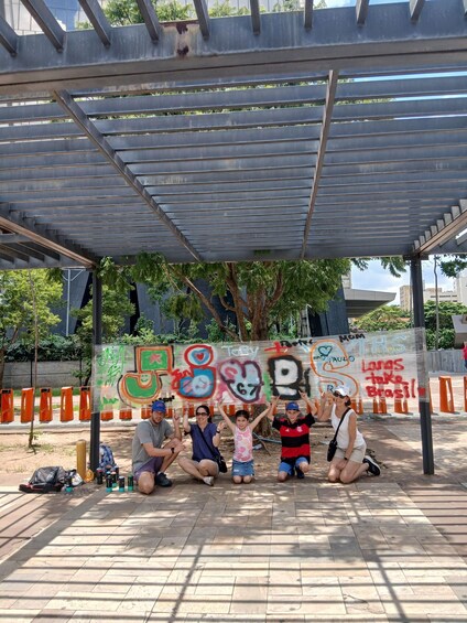 A group of people sitting under a metal structure with graffiti on the wall behind them. 5