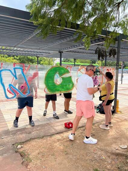 People engaged in an outdoor activity, possibly a sport, under a covered area with trees and a bus in the background. 3