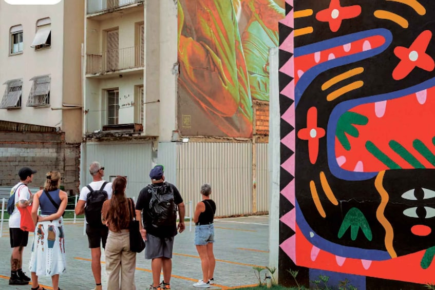 A group of people standing in a parking lot with a colorful mural on the wall behind them. 1