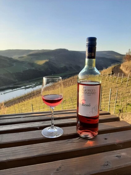 A bottle of Georges Reinhard Rosé wine and a glass of red wine on a wooden bench with a scenic view of hills and a river in the background. 5