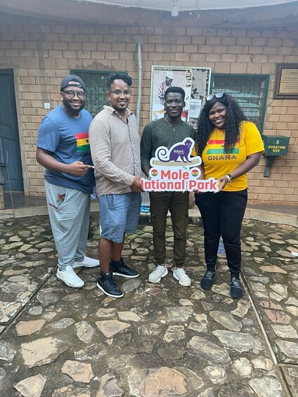 Four people standing in front of a brick building with a donation box and a sign for Mole National Park. 4