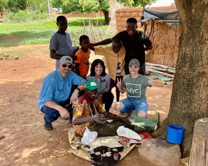 A group of people outdoors, some sitting and others standing, with a tree and a makeshift shelter in the background. 3