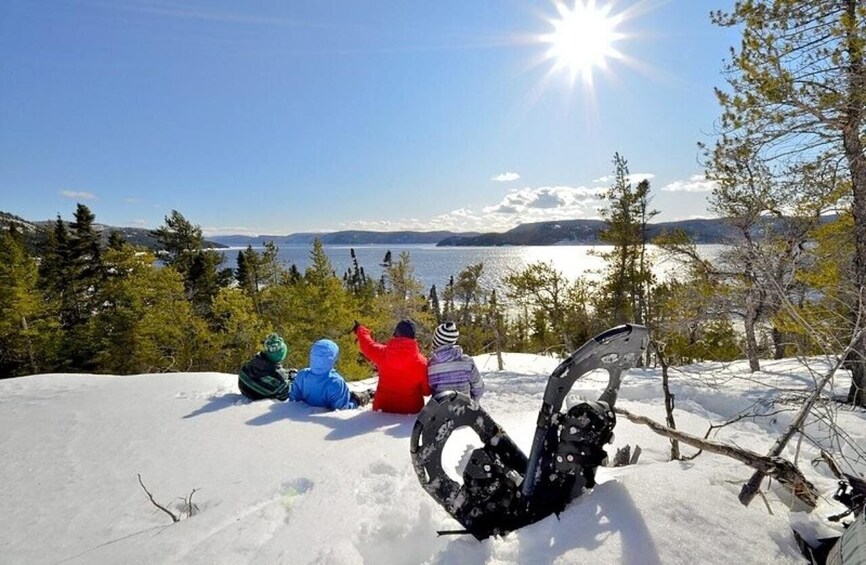 A group of people sitting on a snowy hill overlooking a lake with mountains in the distance. 1
