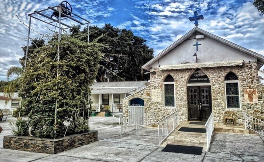 A stone church with a cross on top, a bell tower, and a large evergreen tree in the foreground. 3