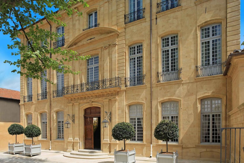 A large, classical building with multiple windows and balconies, surrounded by well-maintained trees and white planters. 1