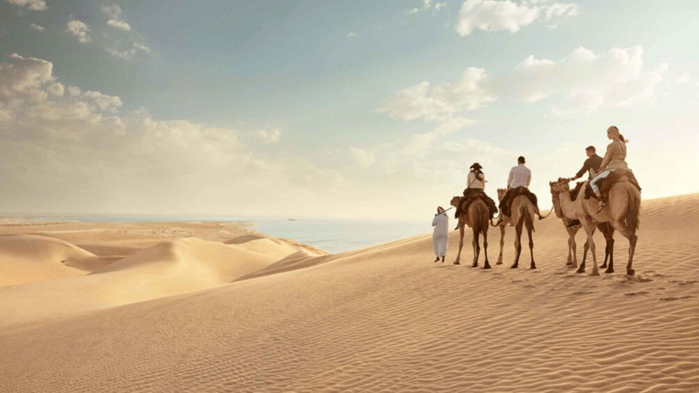 A group of people riding camels across a desert landscape with sand dunes and a body of water in the distance. 3