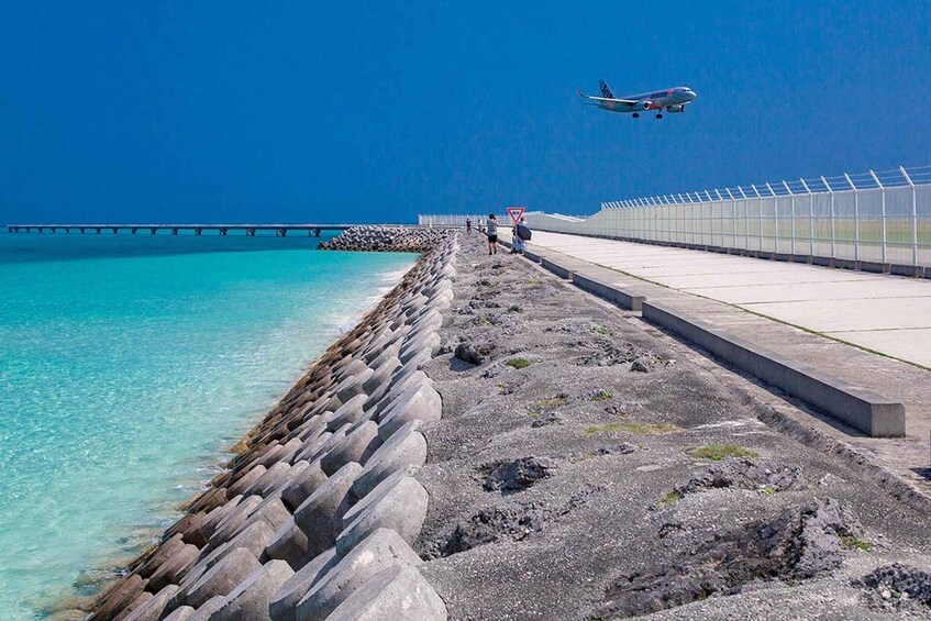 A coastal scene with a clear blue sky, a jet plane flying overhead, a long pier extending into the ocean, and a walkway with a metal fence. 2