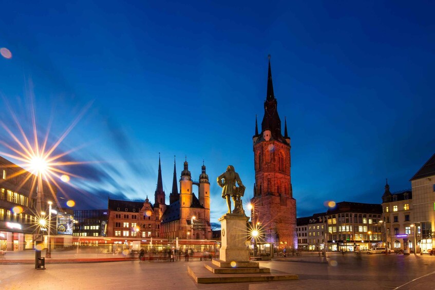 A city square with a prominent clock tower, a statue, and surrounding historic buildings. 2