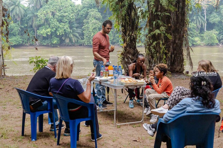 A group of people dining outdoors by a river in a forested area. 2