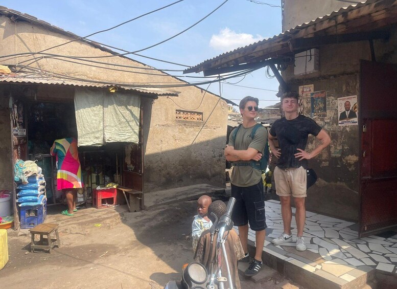 A street scene with a man standing on a tiled platform, another man with a backpack, and a child sitting on the ground. There are buildings with corrugated metal roofs and a sign on the wall. 5