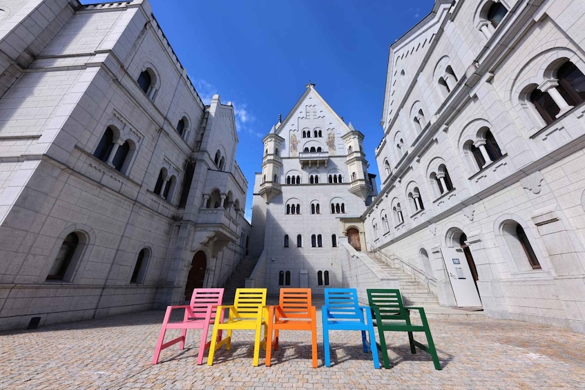 A historic building with arched windows and a central tower. In front, colorful chairs are arranged on a cobblestone ground. 1