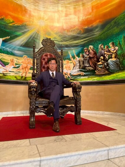 A person seated on an ornate chair with a detailed painting in the background. 3