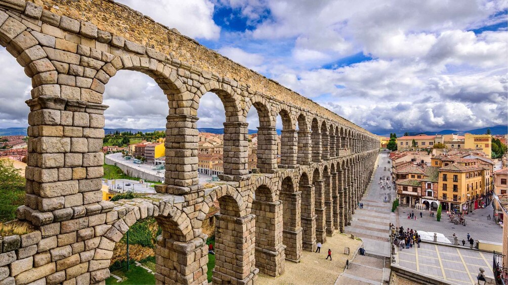 An ancient aqueduct with multiple arches, a paved square with people, and a cityscape in the background. 3