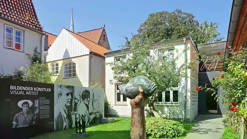 A building with a tiled roof, a garden with a sculpture, and a poster of a visual artist. 2