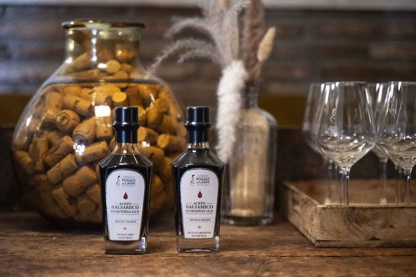 Two bottles of Poggio Al Laghi balsamic vinegar on a wooden surface, with a large jar of olives and two empty wine glasses in the background. 3