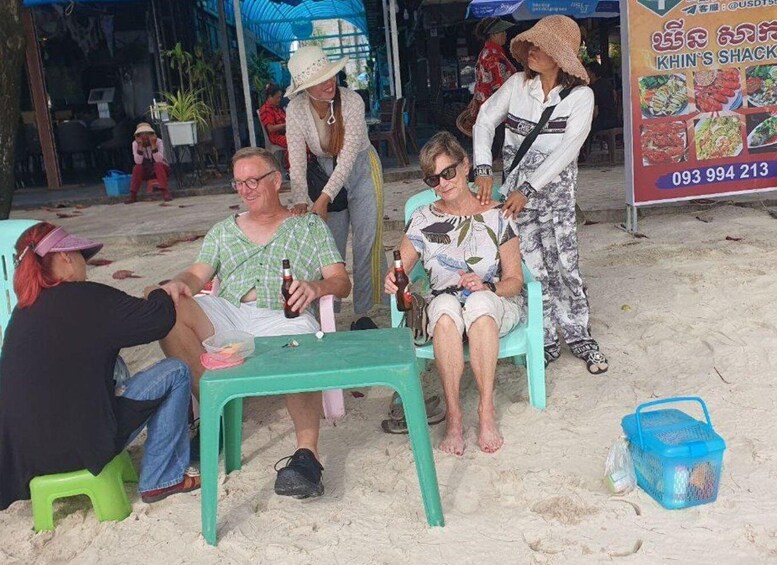 A group of people sitting on plastic chairs on a sandy beach, with a signboard in the background. 3
