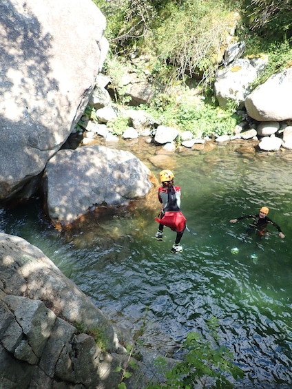 A person in a red and black wetsuit and yellow helmet jumping into a clear water body surrounded by rocks and greenery. 2