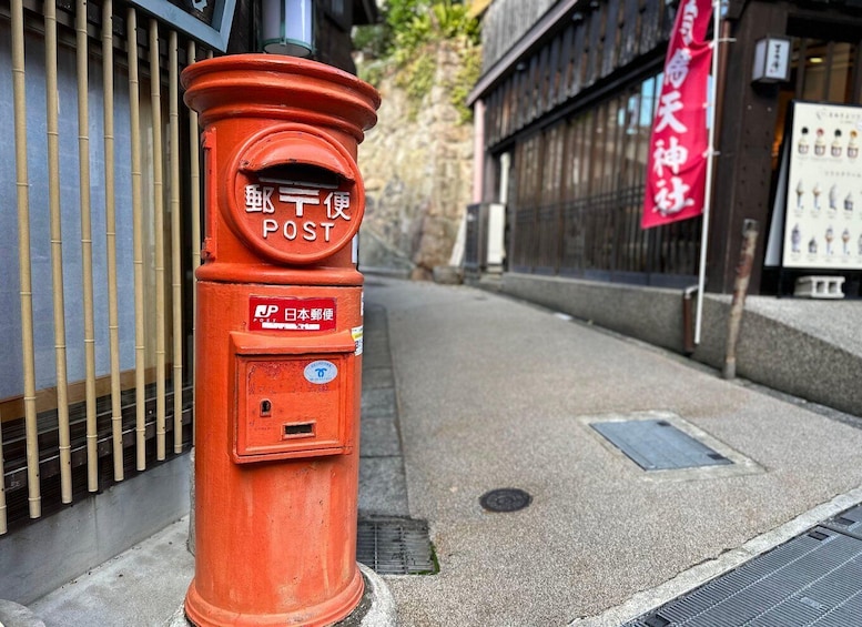 A red Japanese post box on a street with a bamboo fence and a red banner on a building. 5