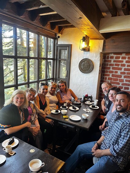 A group of people seated around a table with food and drinks, in a room with wooden beams and a brick wall. 4