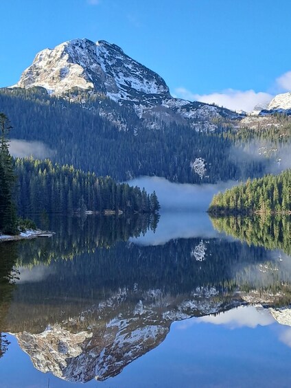 A mountain with snow, a forest, and a calm lake reflecting the scenery. 4