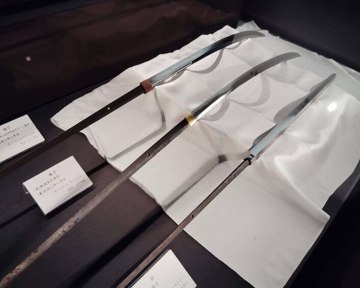 Three swords displayed on a dark surface with white cloths and labels. 3
