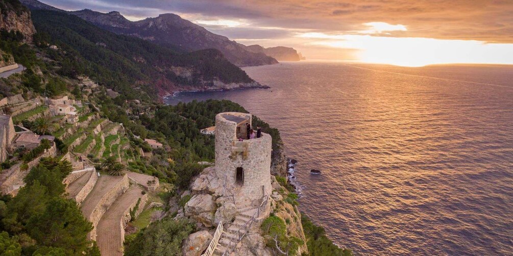 A coastal landscape with terraced fields, a stone tower, and a sunset over the sea. 5