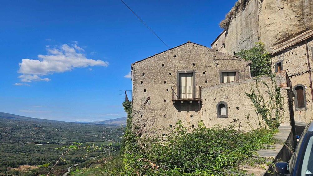 A stone building with a balcony, surrounded by greenery and overlooking a hilly landscape. 1