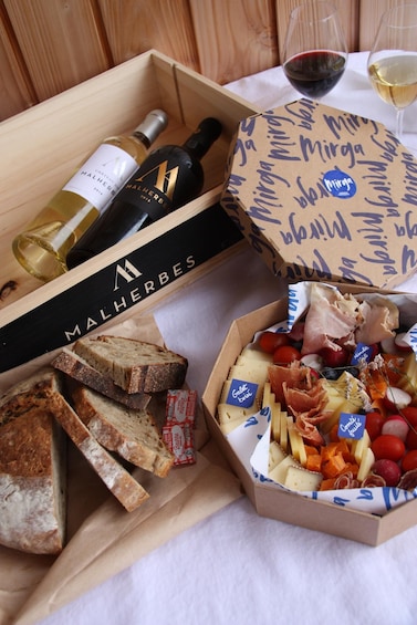 A wooden box with wine bottles, a box of cheese and meats, slices of bread, and a wine glass on a table. 4