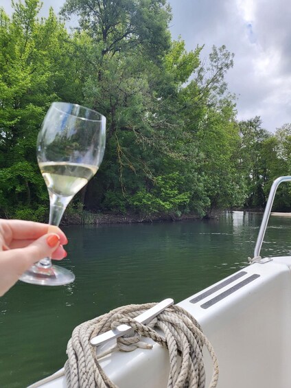 A hand holding a wine glass on a boat with a rope and a water body surrounded by trees. 2