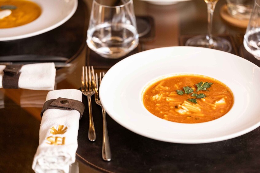 A white plate with a soup garnished with parsley, accompanied by a fork and knife on a table with a napkin and a glass of water. 3