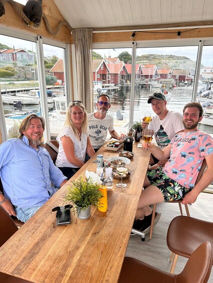 A group of people seated at a wooden table with drinks and snacks, overlooking a marina with boats and buildings. 5