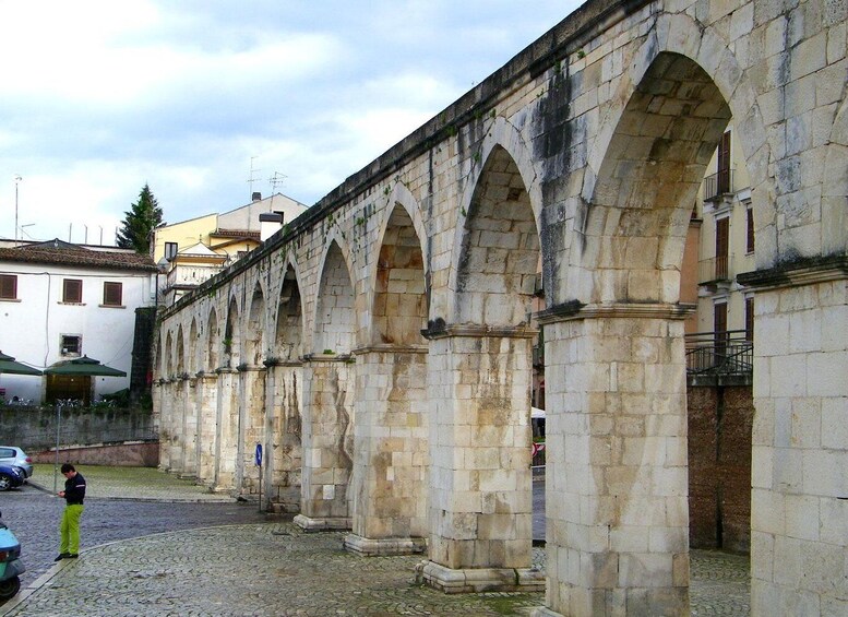 A stone bridge with multiple arches, a cobblestone ground, and buildings in the background. 3