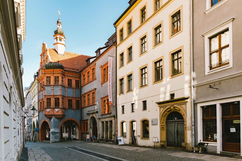 A cobblestone street lined with historic buildings, including a prominent building with a tower and a sign for the Schlesisches Museum zu Görlitz. 1