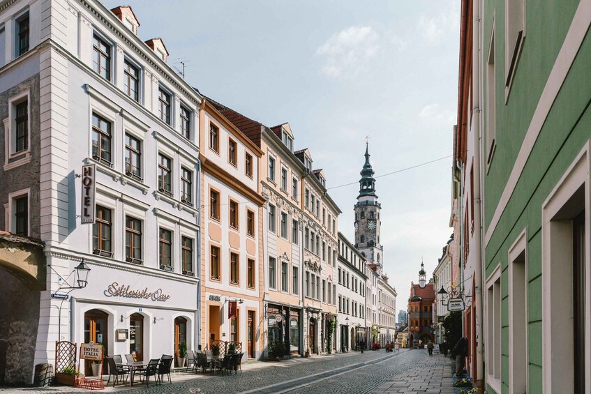 A cobblestone street lined with historic buildings, a prominent clock tower, and outdoor seating at a café. 3