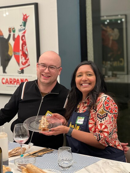 Two people at a dining table with a plate of food, a wine glass, and a poster in the background. 3