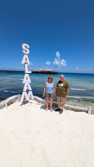 Two people standing on a sandy beach with a sign that reads 'SALAM' and a pier in the background. 2