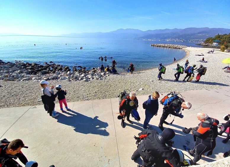 A coastal area with people preparing for water activities, a rocky shoreline, and a clear view of the sea and distant mountains. 4