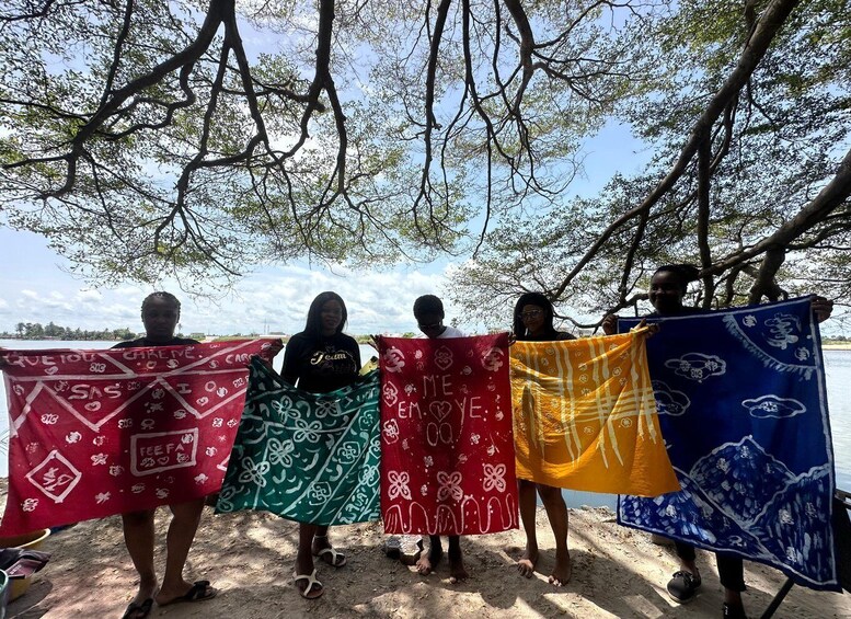 Four individuals holding colorful cloths with various patterns and text, standing under a tree by a body of water. 4