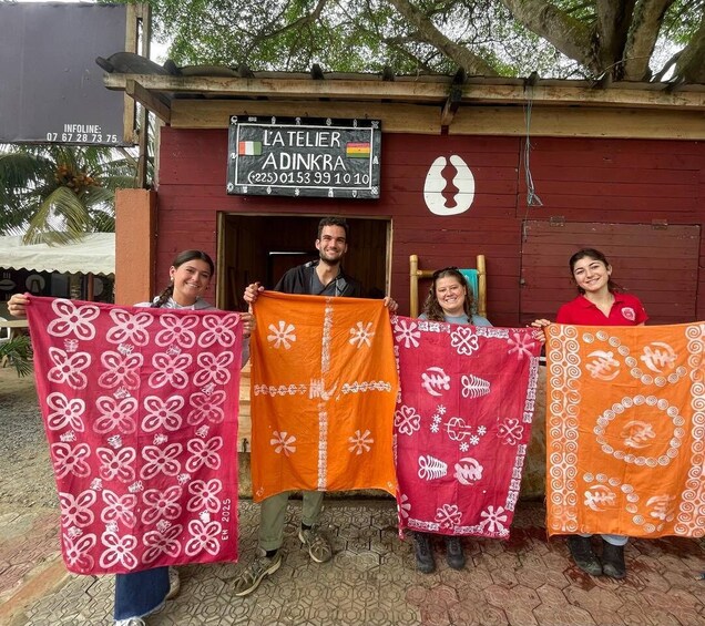 A group of people holding colorful blankets in front of a building with a sign that reads 'L'ATELIER ADINKRA.' 1
