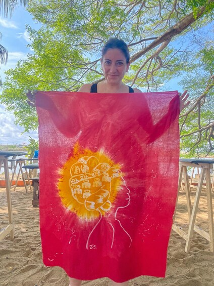 A person holding a red and yellow flag with a lion's head design, standing on a sandy beach with trees in the background. 2