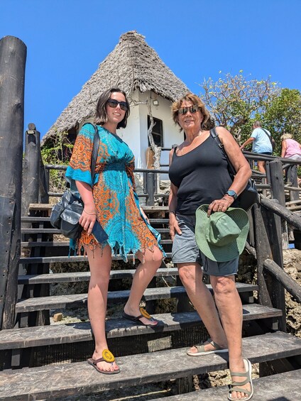 Two people standing on wooden steps with a thatched-roof structure in the background. 1