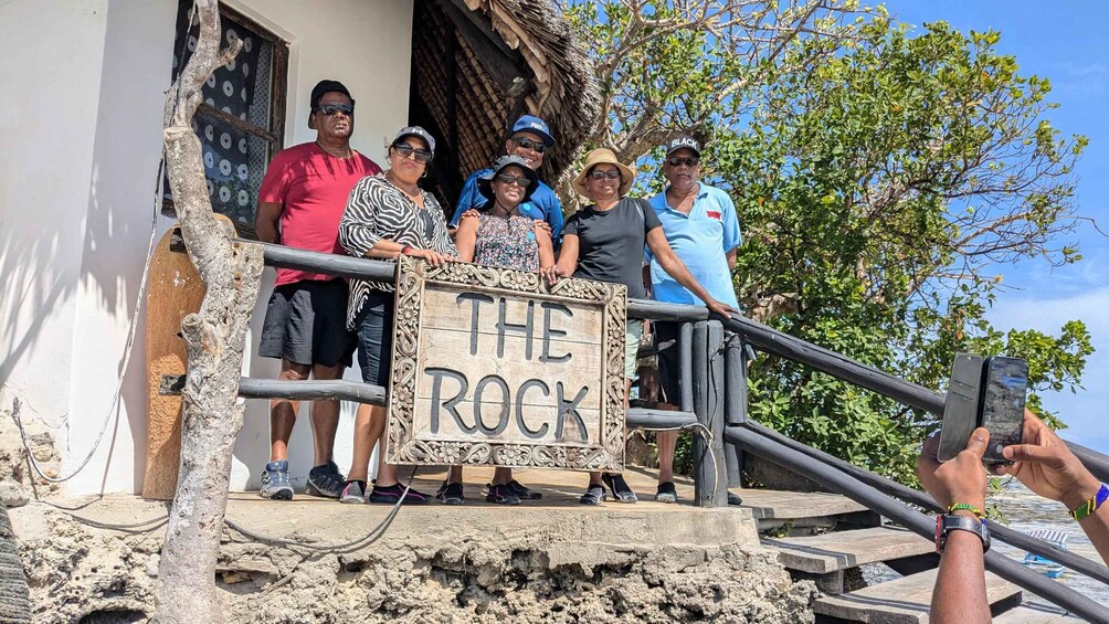 A group of people holding a sign that reads 'THE ROCK' on a staircase with a rustic building in the background. 5