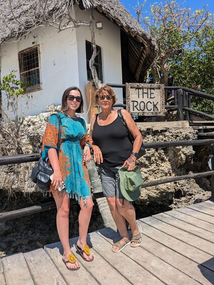 Two people standing on a wooden deck with a sign reading 'THE ROCK' in the background. 2