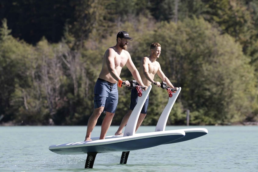Two individuals water skiing on a white board with red handles, surrounded by calm water and a forested shoreline. 1
