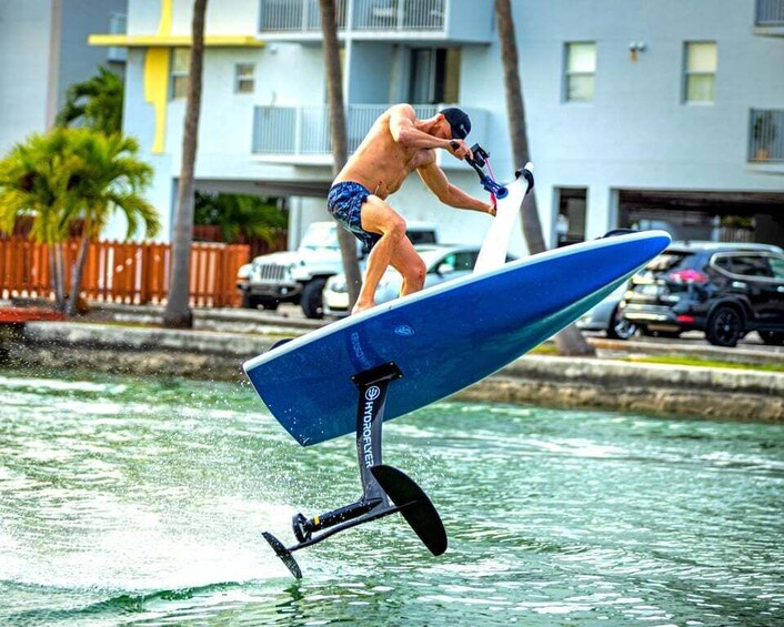 A person riding a hydrofoil board over water, with a residential area and parked cars in the background. 2