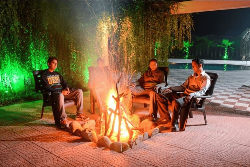 Three people sitting by a campfire in an outdoor setting with greenery and a checkered floor. 5