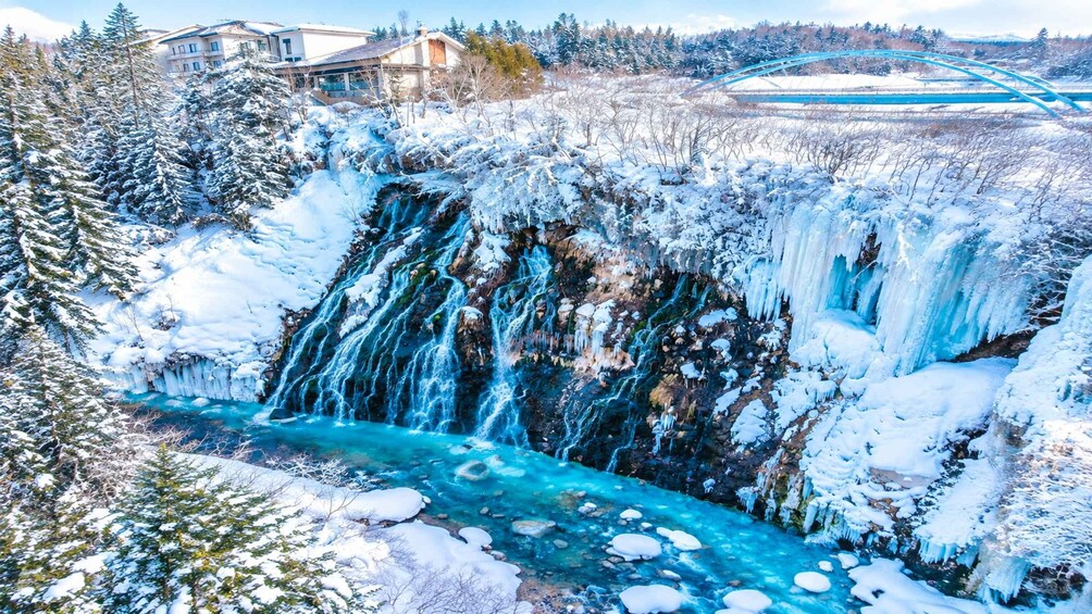 A snow-covered waterfall with a clear blue stream below, surrounded by trees and buildings. 1