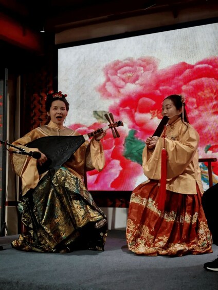 Two performers in traditional attire playing stringed instruments on stage with a floral backdrop. 2